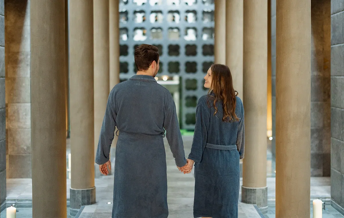 Paar in Bademänteln hält Händchen und geht durch einen stilvollen Spa-Gang mit Säulen und warmem Licht