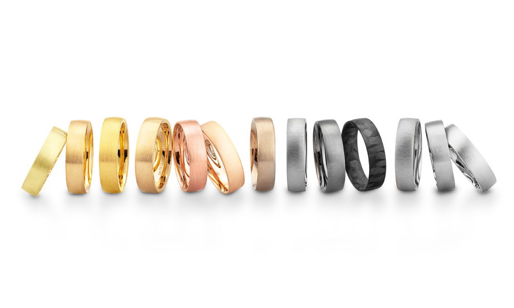 Eine stilvolle Auswahl an Trauringen aus verschiedenen Materialien, darunter warmes Gelbgold, zartes Roségold, kühles Weißgold und markantes Schwarz. Die Bandbreite reicht von schlichten Designs bis hin zu modernen Strukturen mit matten oder gehämmerten Oberflächen.