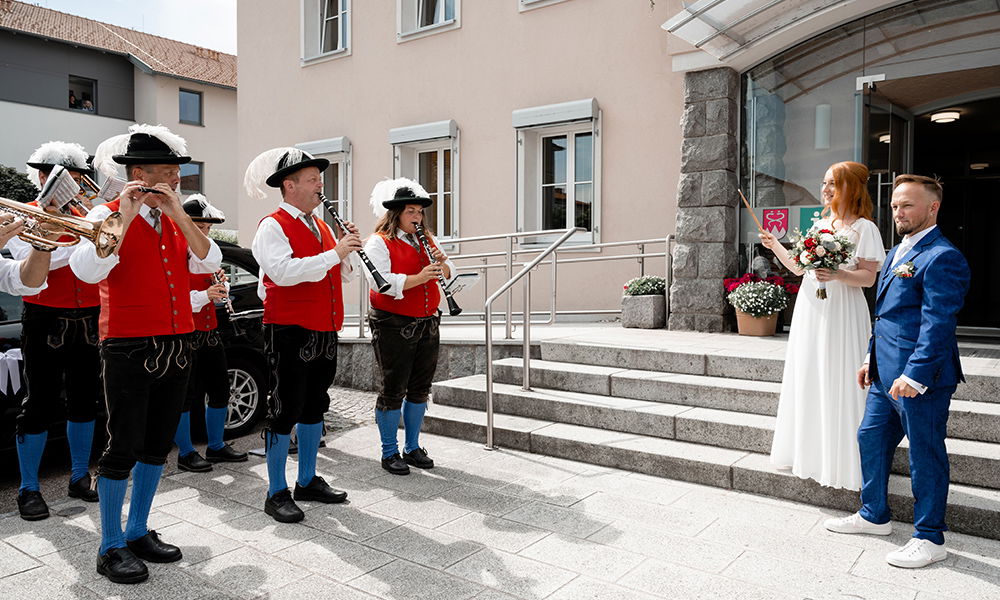 Eine traditionelle Blaskapelle in Tracht spielt vor dem Standesamt, während die Braut Maria mit einem Hochzeitsstab und Bräutigam Markus in blauem Anzug die musikalische Darbietung genießen.