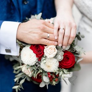 Die frisch vermählten Hände von Maria und Markus mit ihren Eheringen ruhen auf einem Brautstrauß aus roten und weißen Rosen, umgeben von grünen Blättern und dezenten Beeren.