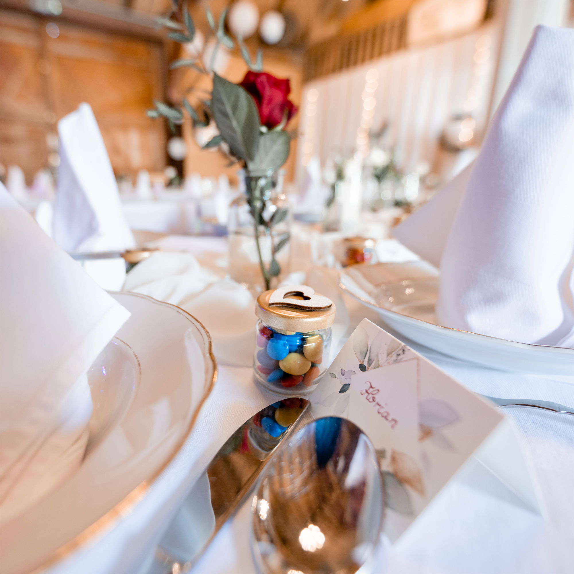 Eine elegant gedeckte Hochzeitstafel mit weißen Servietten, goldgerandetem Geschirr und einem kleinen Gastgeschenk in Form eines Gläschens mit bunten Schokolinsen. Eine rote Rose und eine Namenskarte schmücken den Platz.