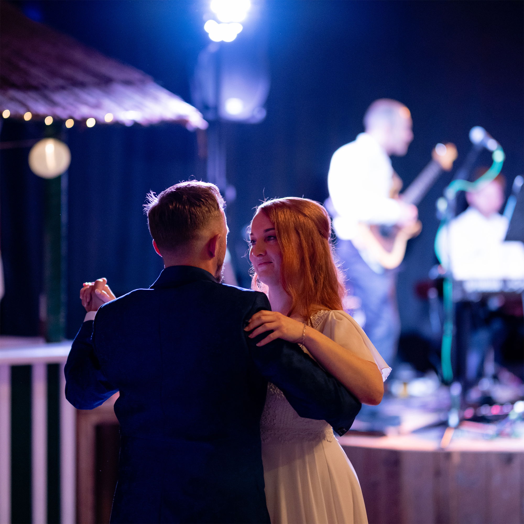 Maria und Markus tanzen eng umschlungen auf der Hochzeitsfeier. Im Hintergrund spielt eine Live-Band, während die Braut ihren frisch angetrauten Ehemann liebevoll ansieht.