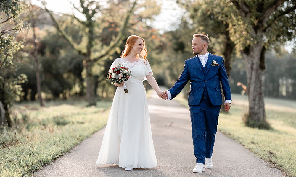 Maria und Markus spazieren Hand in Hand über einen idyllischen Weg in der Natur. Die Braut trägt ein schlichtes, elegantes Kleid, der Bräutigam einen blauen Hochzeitsanzug mit floralem Muster.
