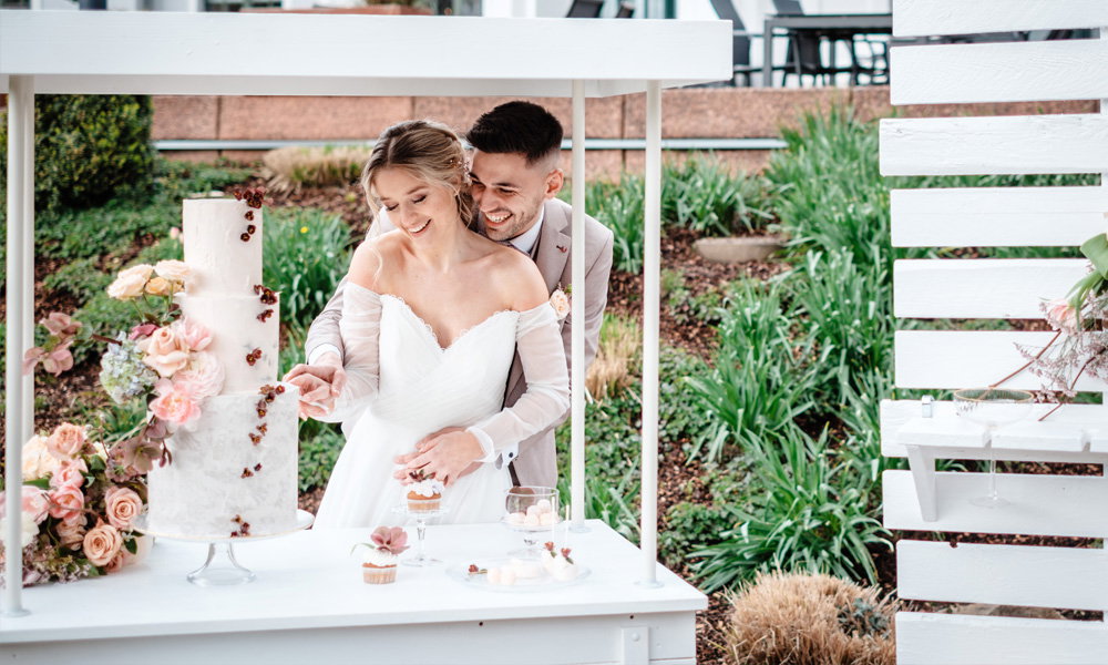 Romantischer Moment: Braut und Bräutigam lächeln beim Anschneiden der Torte, dekoriert mit Blumen und Beeren, inmitten eines grünen Gartens.