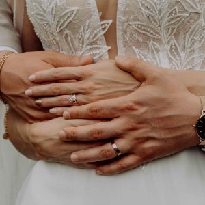 Nahaufnahme der Hände eines frisch verheirateten Paares: Der Bräutigam hält die Braut liebevoll umarmt, während ihre Ringe im Fokus stehen.