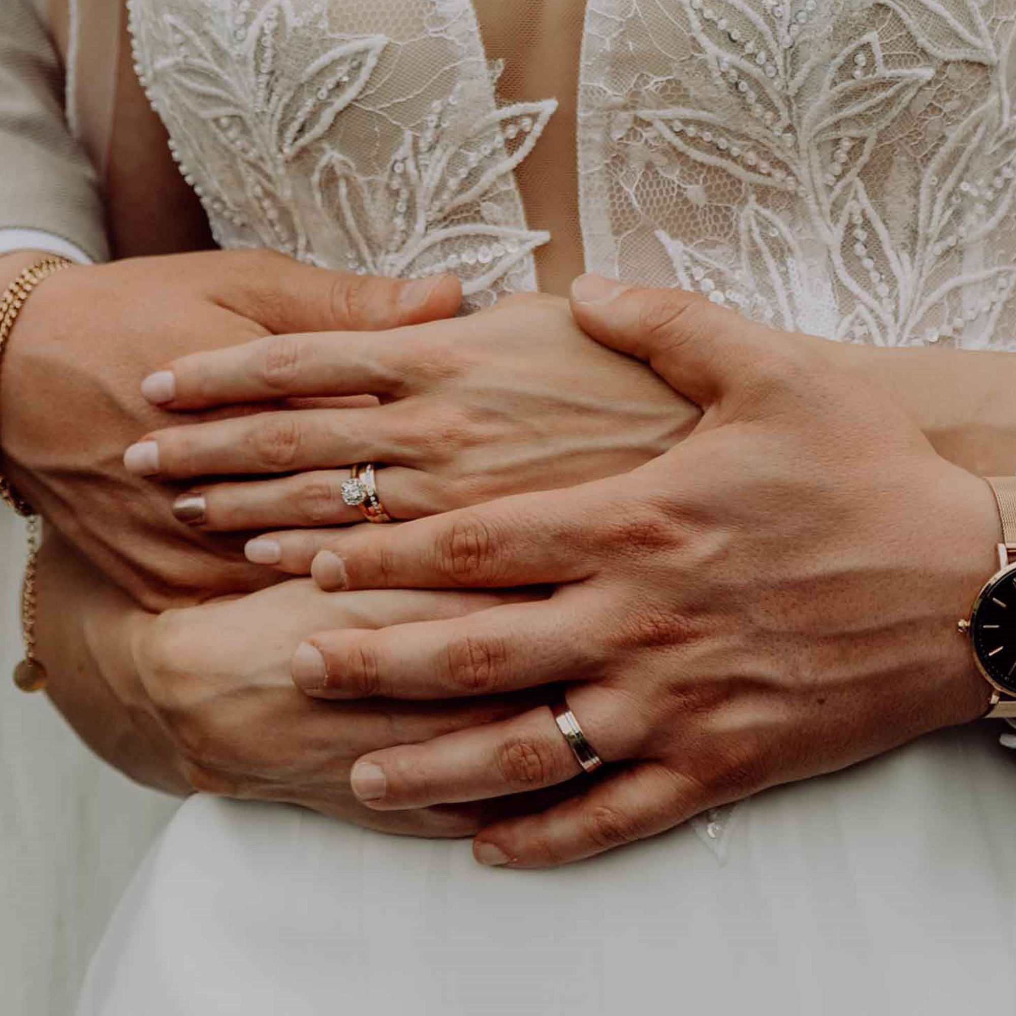 Nahaufnahme der Hände eines frisch verheirateten Paares: Der Bräutigam hält die Braut liebevoll umarmt, während ihre Ringe im Fokus stehen.