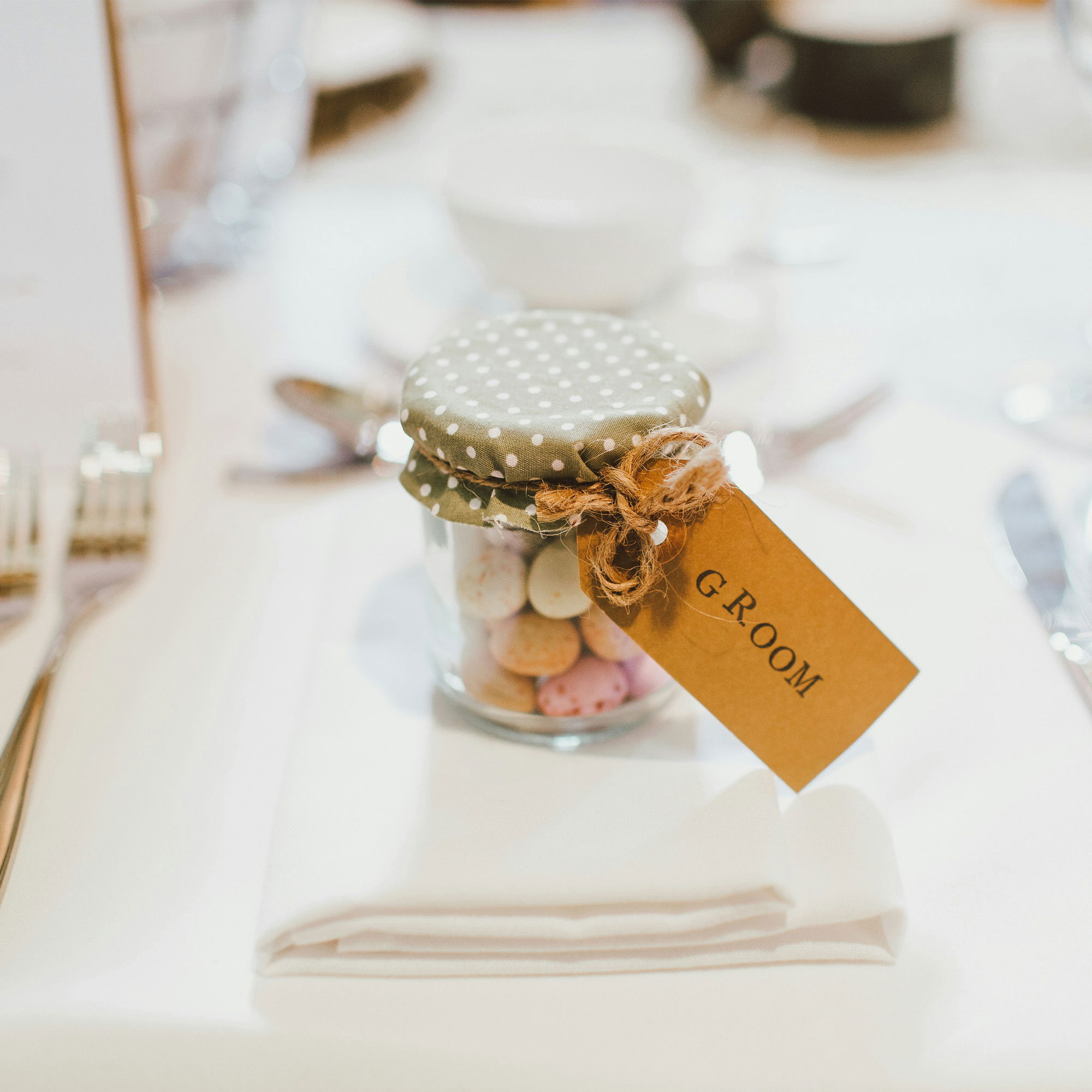 Gastgeschenk aus Glas, gefüllt mit bunten Bonbons, abgedeckt mit gepunktetem Stoff und einer rustikalen Juteschnur, versehen mit einem Anhänger ‚Groom‘.