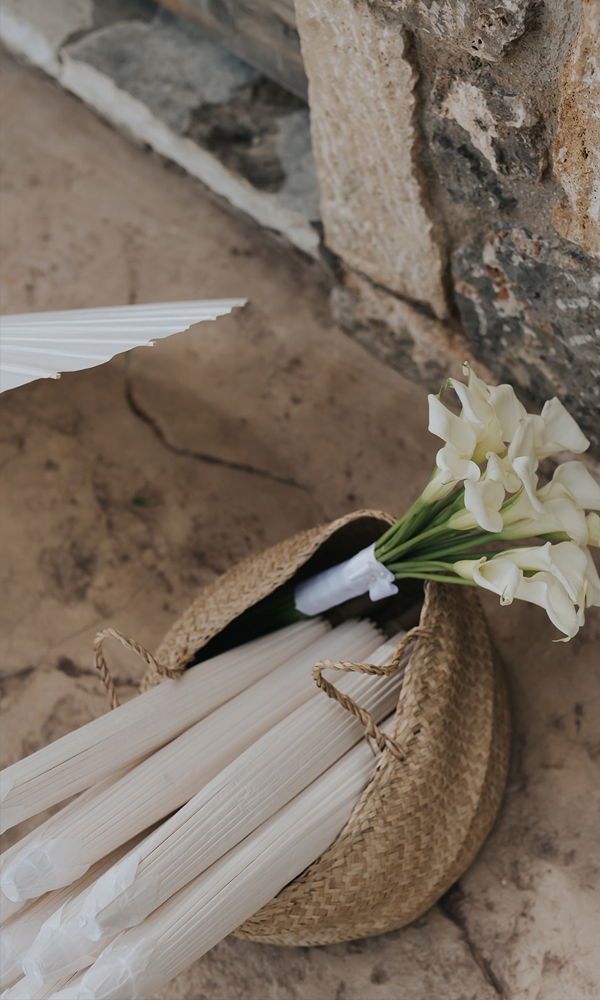 Geflochtene Tasche mit weißen Sonnenschirmen und einem Brautstrauß aus Calla-Blumen – stilvolles Hochzeitsdetail in mediterraner Umgebung.