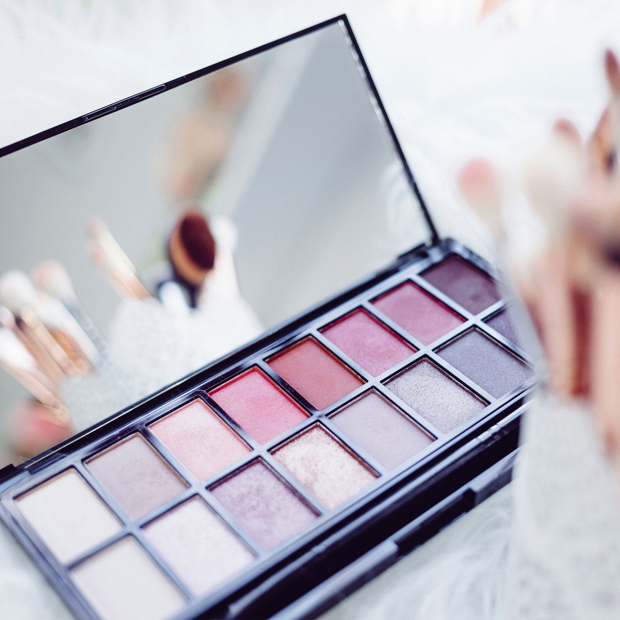 Beauty-Flatlay: Eine geöffnete Lidschattenpalette mit verschiedenen Nude- und Rottönen, im Hintergrund verschwommene Make-up-Pinsel.