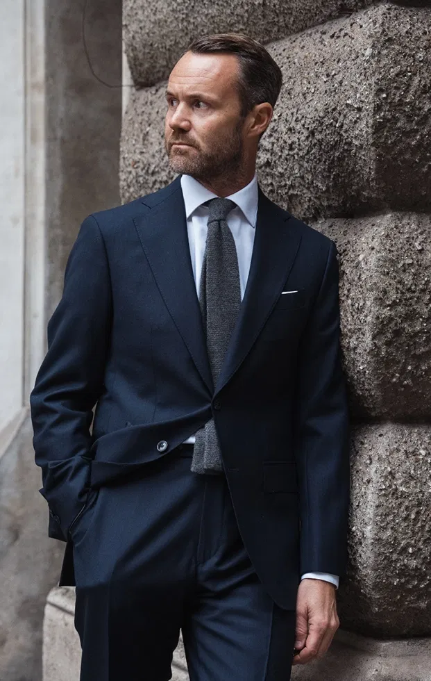 Elegant gekleideter Mann in dunklem Businessanzug lehnt an einer Steinwand und blickt zur Seite