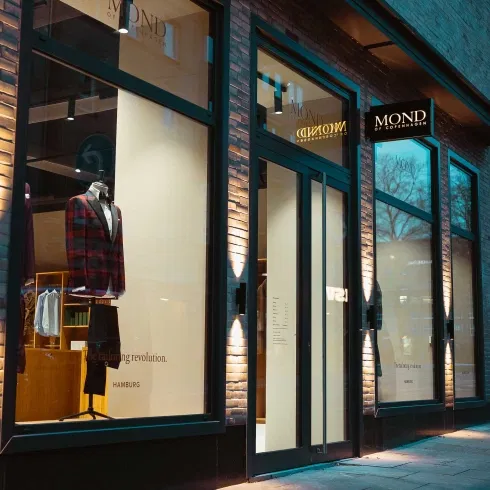 Beleuchtetes Schaufenster eines Herrenausstatters mit elegantem Anzug auf einer Schneiderpuppe und moderner Glasfassade in der Abenddämmerung.