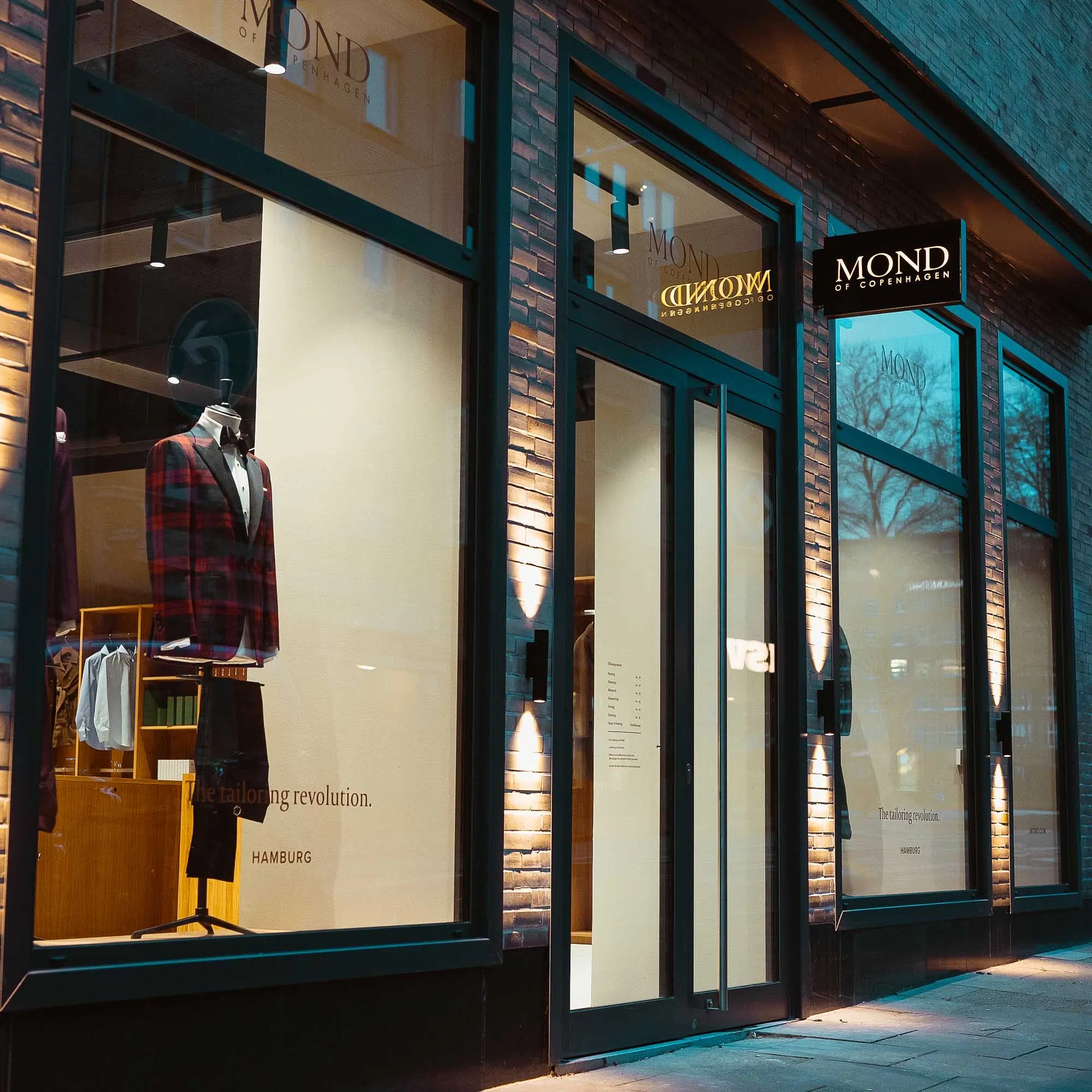 Beleuchtetes Schaufenster eines Herrenausstatters mit elegantem Anzug auf einer Schneiderpuppe und moderner Glasfassade in der Abenddämmerung.