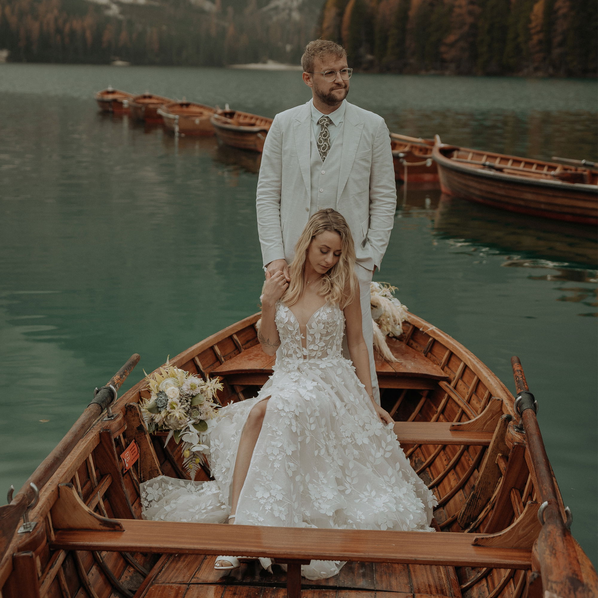 Romantische Hochzeitsaufnahme: Ein Bräutigam in einem cremefarbenen Anzug steht aufrecht im Boot und hält die Schulter seiner Braut, die in einem Spitzenkleid verträumt nach unten blickt. Im Hintergrund spiegeln sich herbstliche Bäume im See.