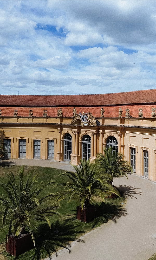 Gelbliches Schloss mit rotem Dach und Palmen im Vordergrund bei blauem Himmel.