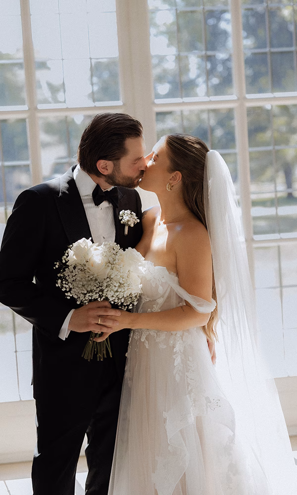 Romantisches Paarfoto am Hochzeitstag mit Brautstrauß und Fenster im Hintergrund.