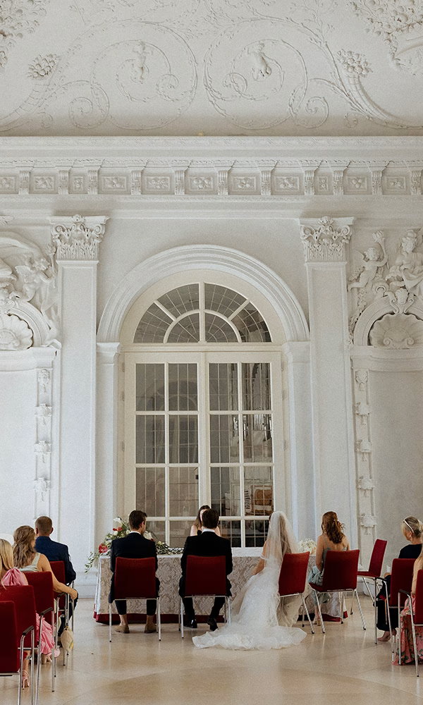 Gäste verfolgen die Zeremonie in einem eleganten Raum mit hohen Fenstern.