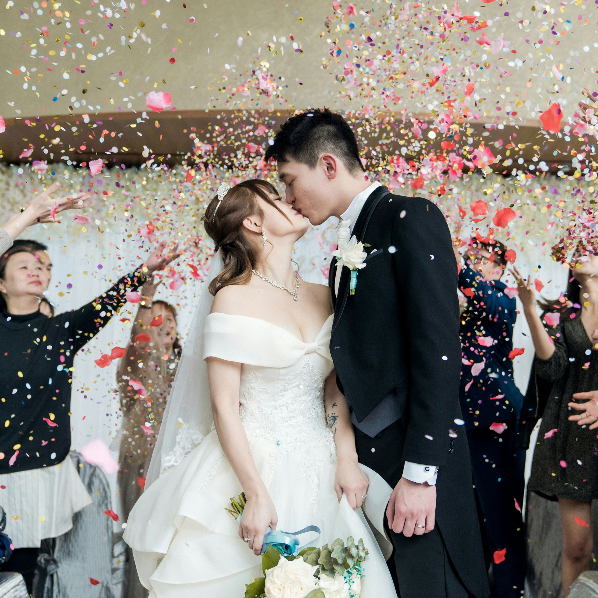 Frisch verheiratetes Paar küsst sich nach der Trauung, während Gäste Konfetti und Blütenblätter werfen.