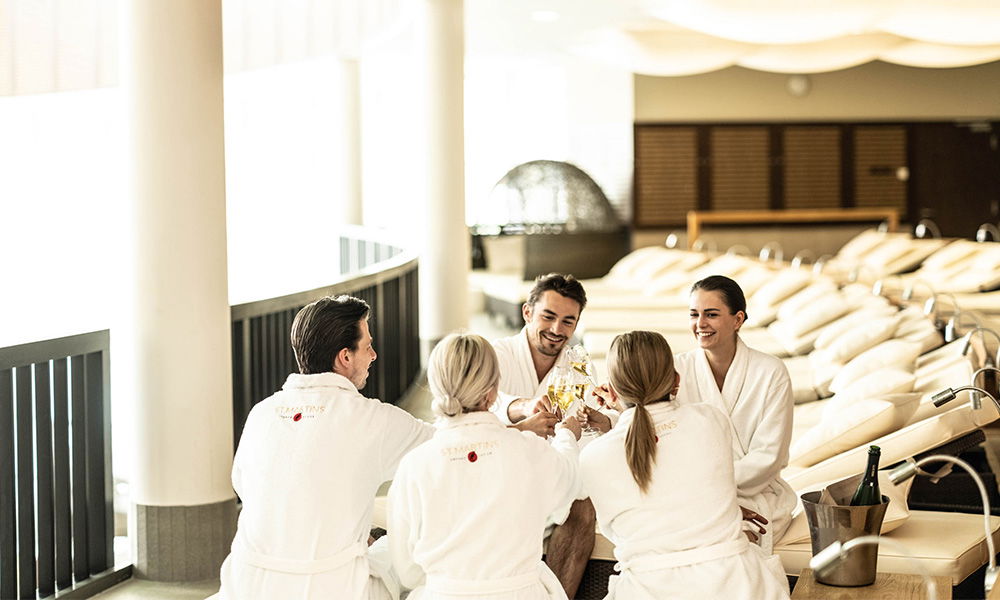 Freunde stoßen im Wellnessbereich in Bademänteln mit Sektgläsern an.
