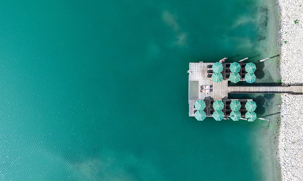 Luftaufnahme eines Holzstegs mit Sonnenliegen und Schirmen am See.