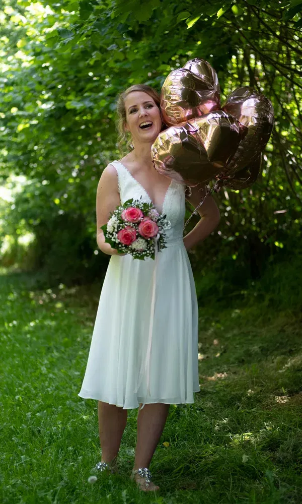 Lachende Braut hält roségoldene Herzluftballons und pinke Blumen in einem sommerlichen Garten.
