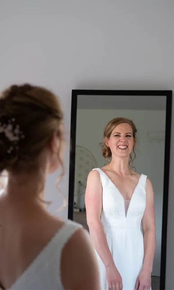 Junge Frau in Brautkleid steht vor einem Spiegel und lächelt glücklich über ihren Anblick.