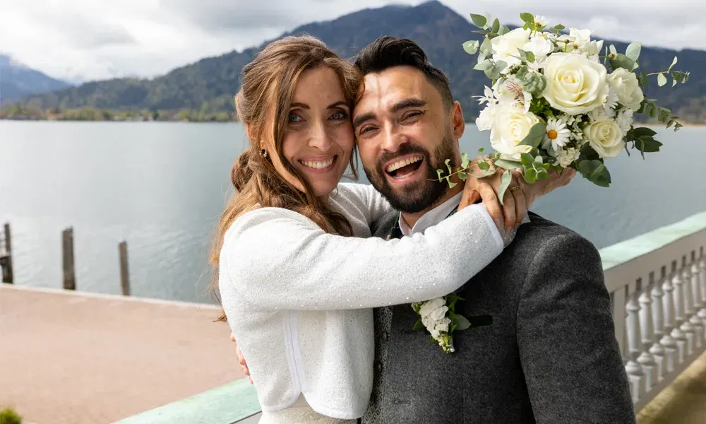 Janine und Martin feiern ihre Trauung am Tegernsee mit einer innigen Umarmung und Blick in die Kamera, der Strauß aus weißen Rosen im Vordergrund.