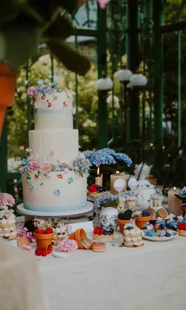 Dreistöckige weiße Torte mit Blüten, umgeben von Gebäck, Beeren und Kerzen auf Hochzeitstisch.