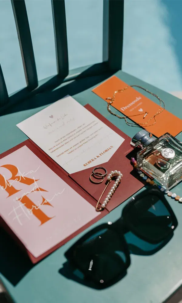 Detailaufnahme von Einladungskarten, Schmuck, Parfumflakon und Sonnenbrille – liebevoll arrangiert für das Hochzeits-Wochenende.