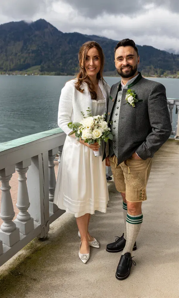 Hochzeitspaar in Dirndl und Lederhose posiert auf einer Terrasse mit Seeblick – traditionelle Hochzeit in malerischer Bergkulisse.