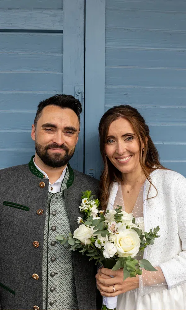 Hochzeitspaar im Dirndl und in Lederhose steht auf einem Balkon mit Seeblick – traditionelle Hochzeit mit Alpenkulisse.
