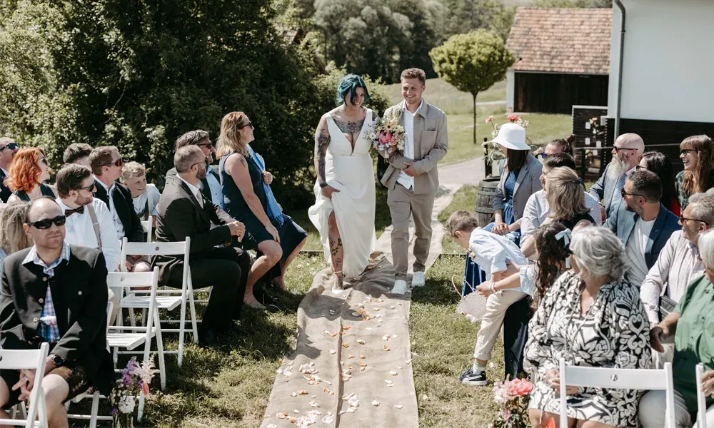 Während der freien Trauung geht die Braut barfuß und lächelnd mit ihrem Sohn durch den Blütenweg, die Gäste blicken erwartungsvoll.