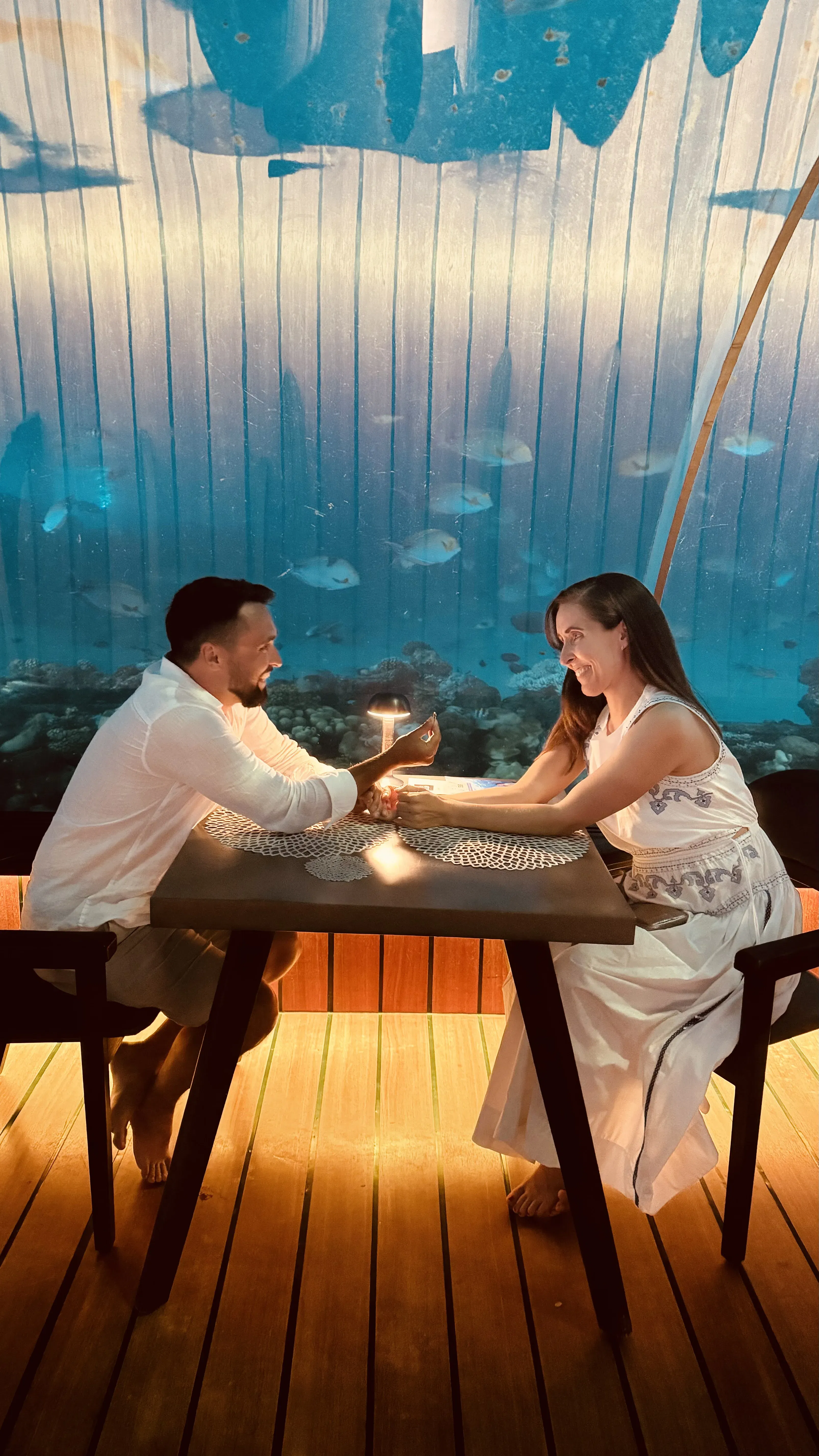 Ein Paar bei einem besonderen Moment im 5.8 Undersea Restaurant – er hält ihre Hand, während Fische im Hintergrund vorbeischwimmen.