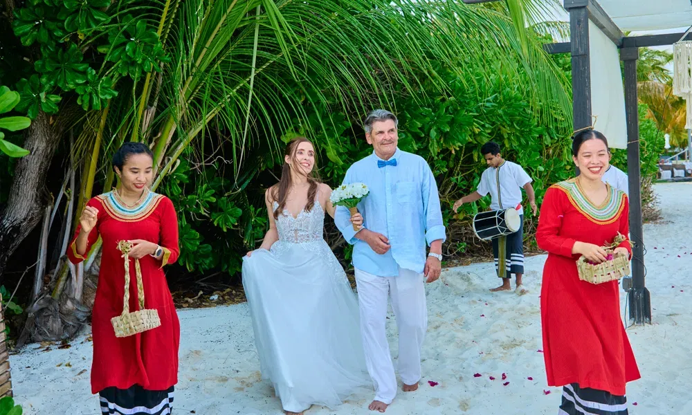 Janine schreitet im Brautkleid barfuß mit ihrem Vater durch den Sand, umgeben von feierlicher Begleitung in roten Gewändern.