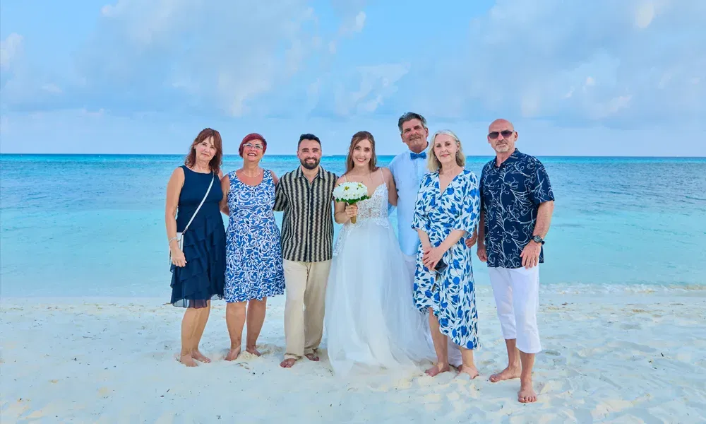 Hochzeitsgesellschaft in sommerlicher Kleidung posiert gemeinsam mit dem Brautpaar am Strand unter blauem Himmel.