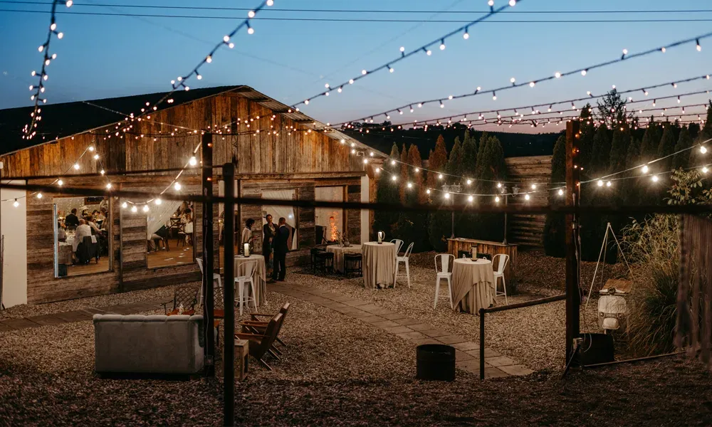Die Outdoor-Location erstrahlt bei Dämmerung in warmem Lichterglanz – Holzhütte, Steinterrasse und Stehtische laden zum Verweilen ein.