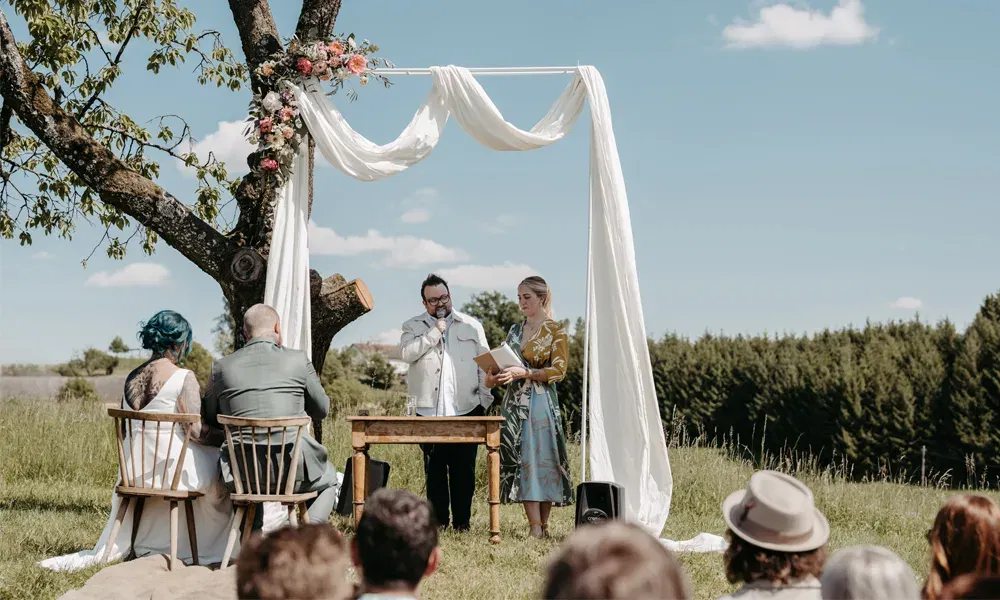 Rebeka und Florian lauschen der Zeremonie auf einer Wiese mit Blumenbogen, Baum und Rednerpaar im Zentrum des Geschehens.