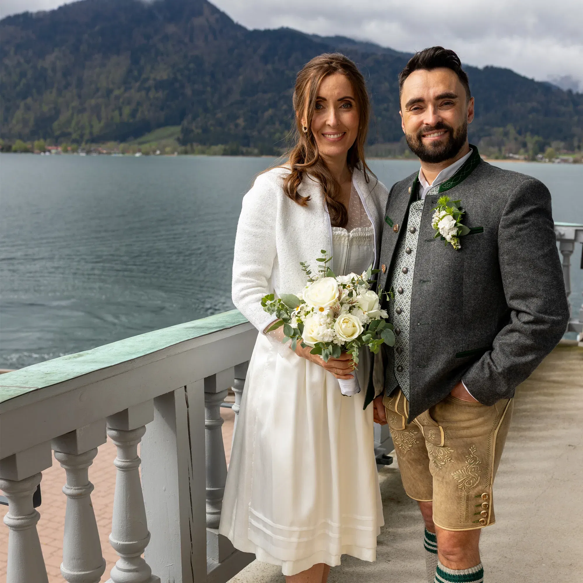 Hochzeitspaar in Dirndl und Lederhose posiert auf einer Terrasse mit Seeblick – traditionelle Hochzeit in malerischer Bergkulisse.
