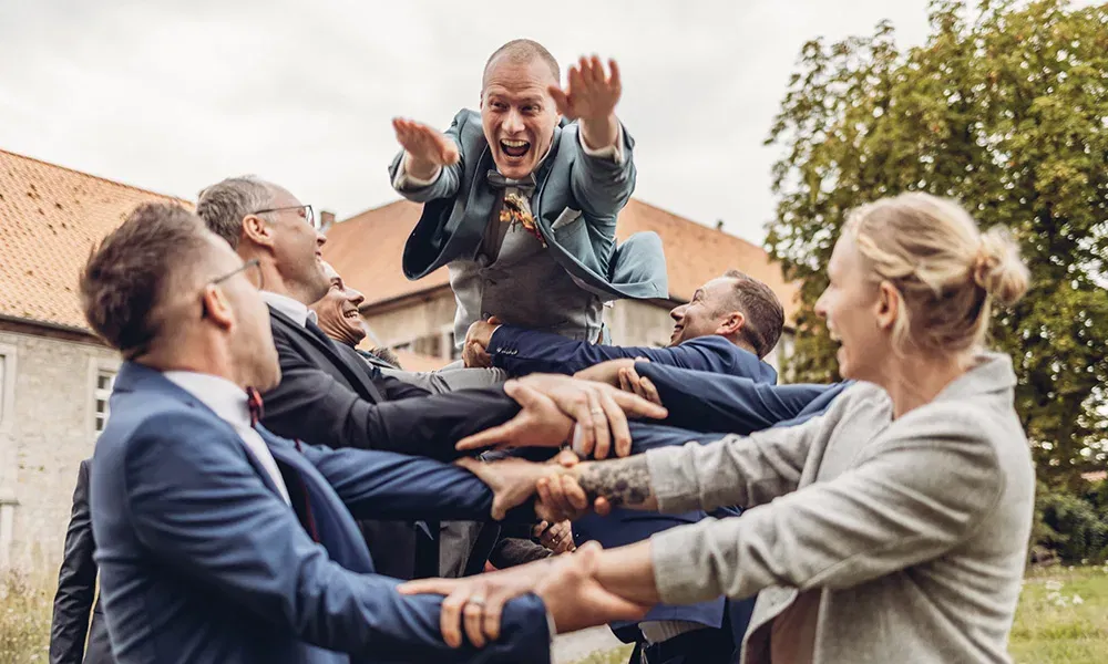 Freundesgruppe wirft den Bräutigam lachend in die Luft bei der Hochzeit.