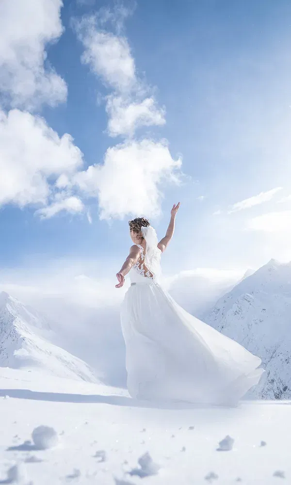 Braut in weißem Kleid tanzt anmutig auf schneebedecktem Berggipfel.