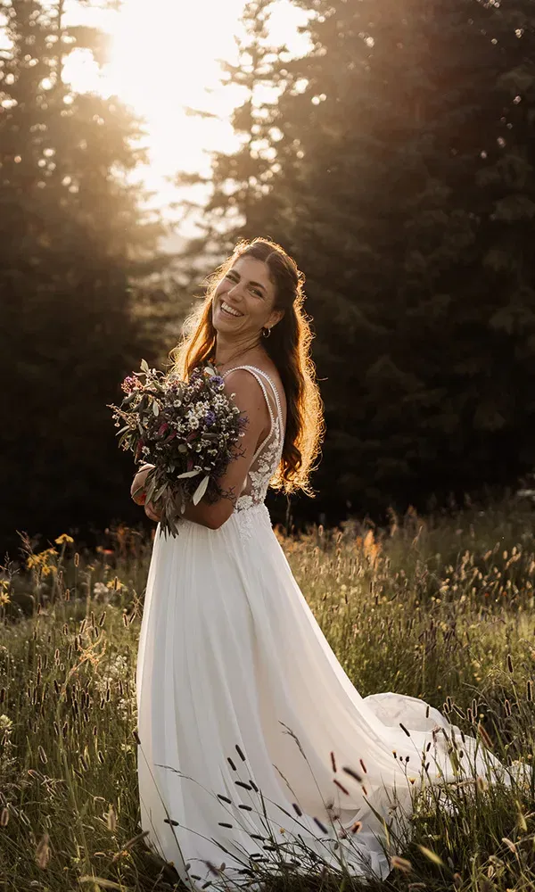 Strahlende Braut mit Blumenstrauß im Gegenlicht einer sommerlichen Wiese.