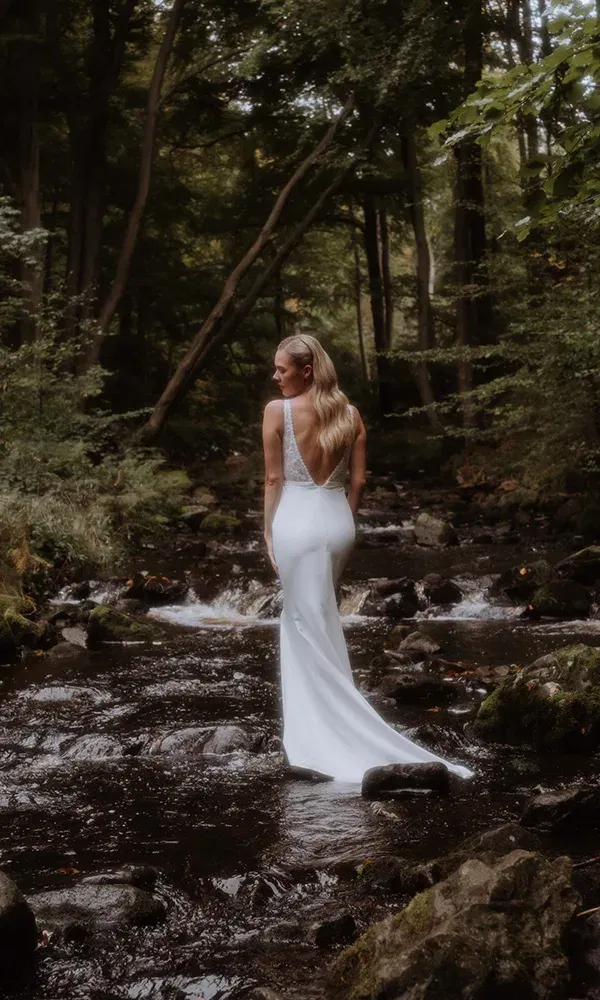 Braut im schlichten Kleid steht verträumt im Bachbett eines Waldes.