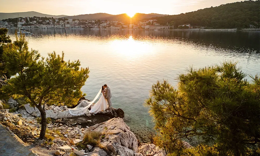 Brautpaar küsst sich auf einer Felsklippe mit Blick auf das Meer bei Sonnenuntergang.
