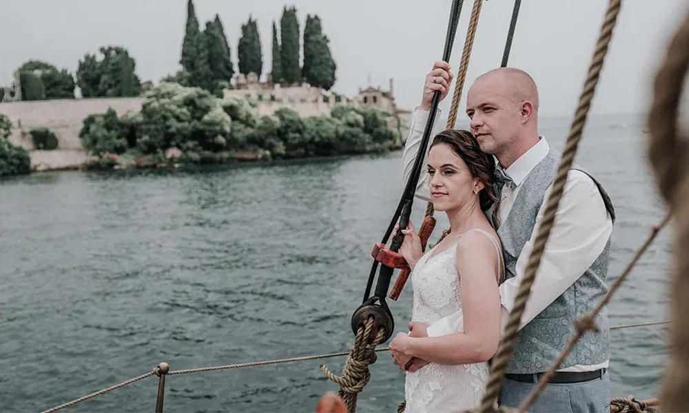 Brautpaar steht verliebt an der Reling eines Segelschiffs mit Blick auf eine Insel.