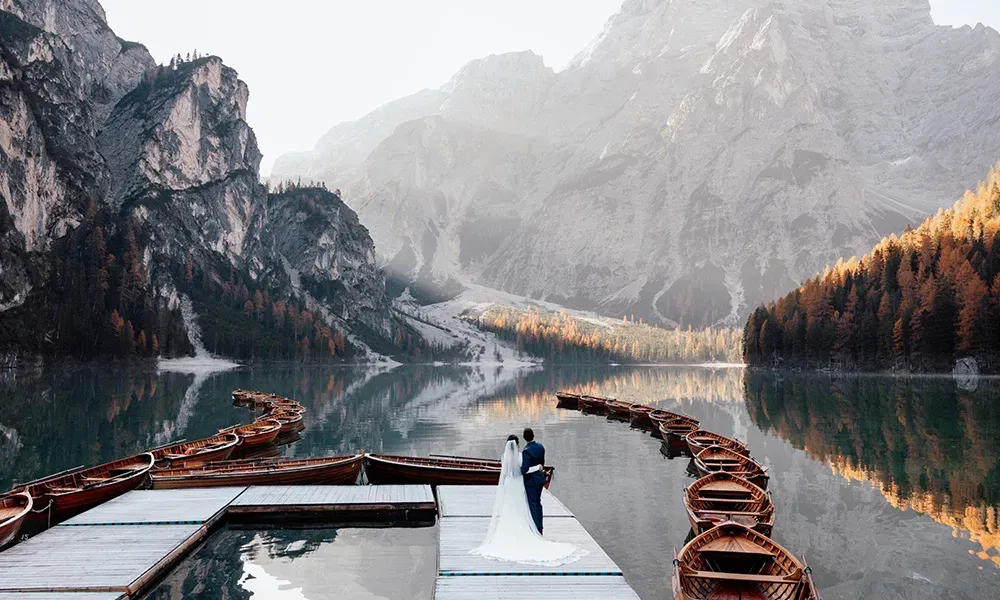 Braut und Bräutigam stehen auf einem Steg am See, umgeben von Booten und Bergen.