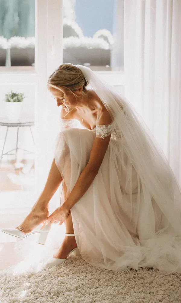 Braut sitzt im Licht und schließt den Riemen ihrer Hochzeitsschuhe.