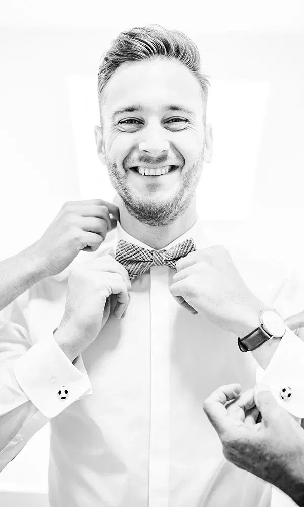 Fröhlicher Bräutigam beim Anziehen seiner Fliege, umgeben von helfenden Händen.