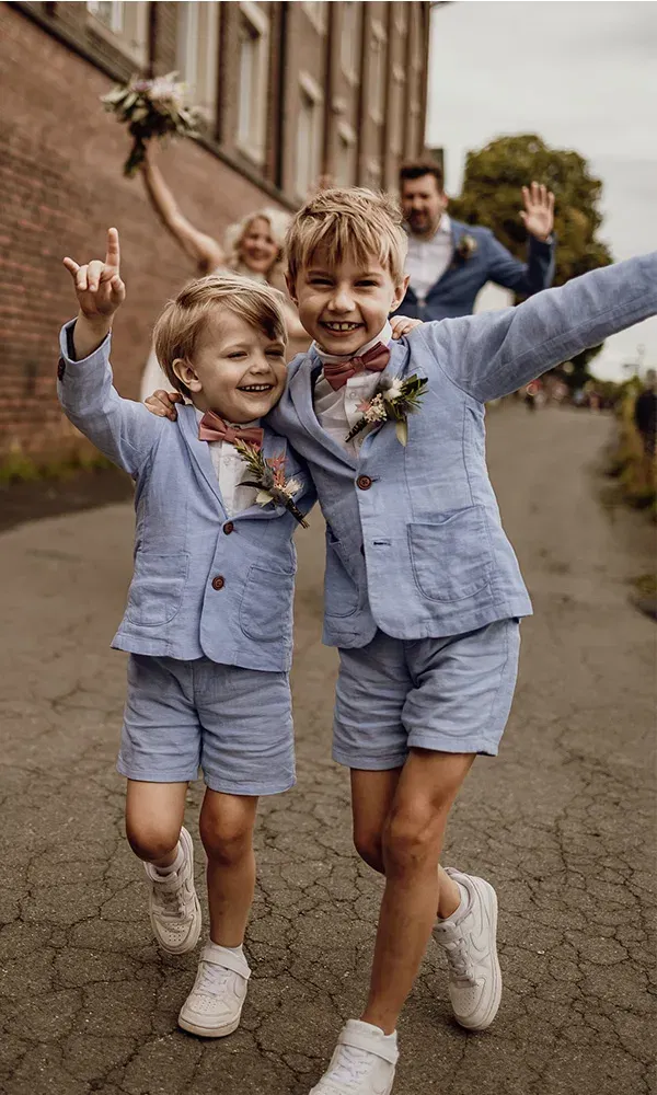 Zwei Jungs in blauen Anzügen laufen lachend auf einer Straße, im Hintergrund jubelt die Familie.