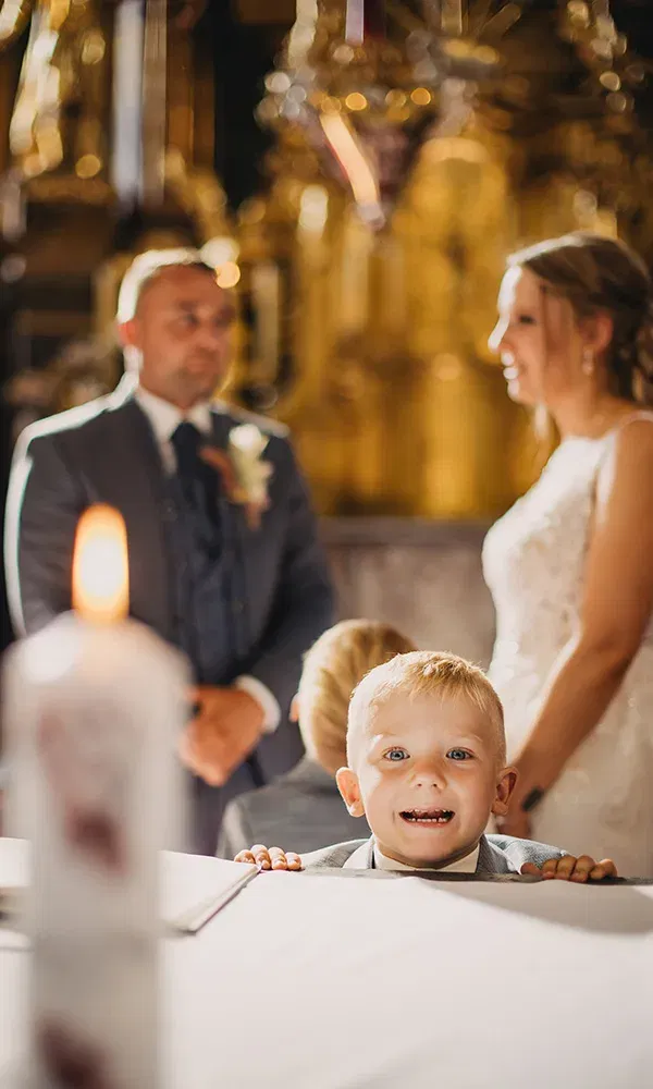 Kleiner Junge blickt neugierig über den Tisch, während das Brautpaar im Hintergrund verschwimmt.