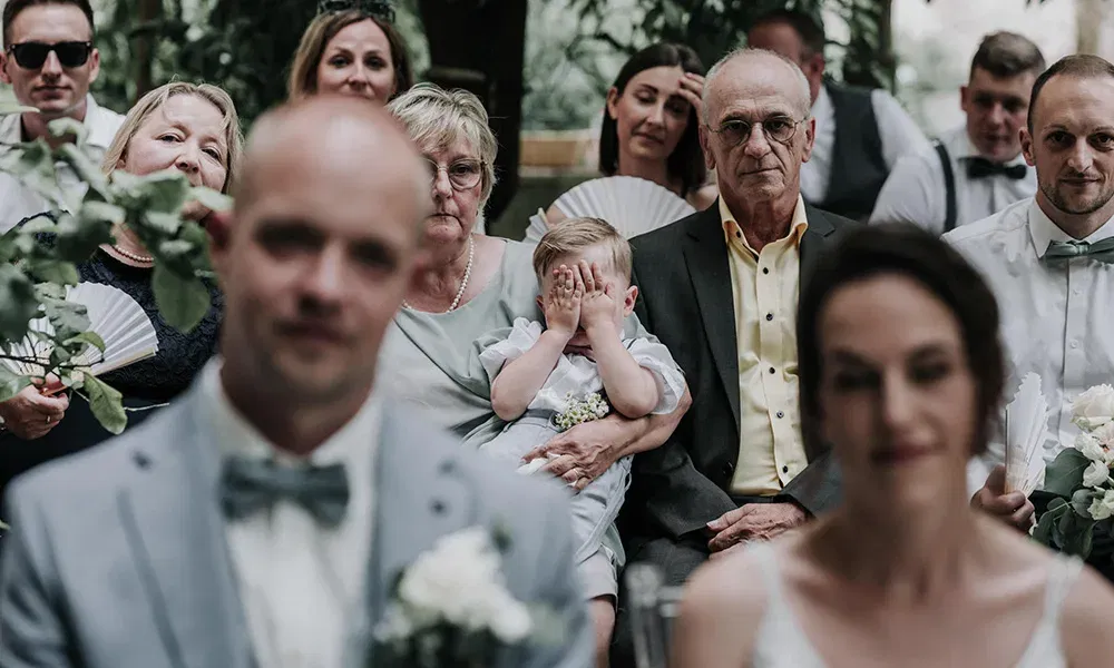 Kleines Kind schämt sich oder spielt, während es sich die Augen zuhält – mitten in der Hochzeitsgesellschaft.