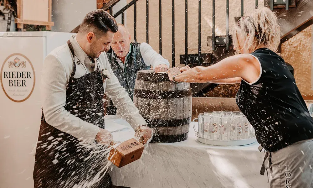 Der Bräutigam schlägt ein Bierfass an, während das Bier in hohem Bogen herausspritzt.